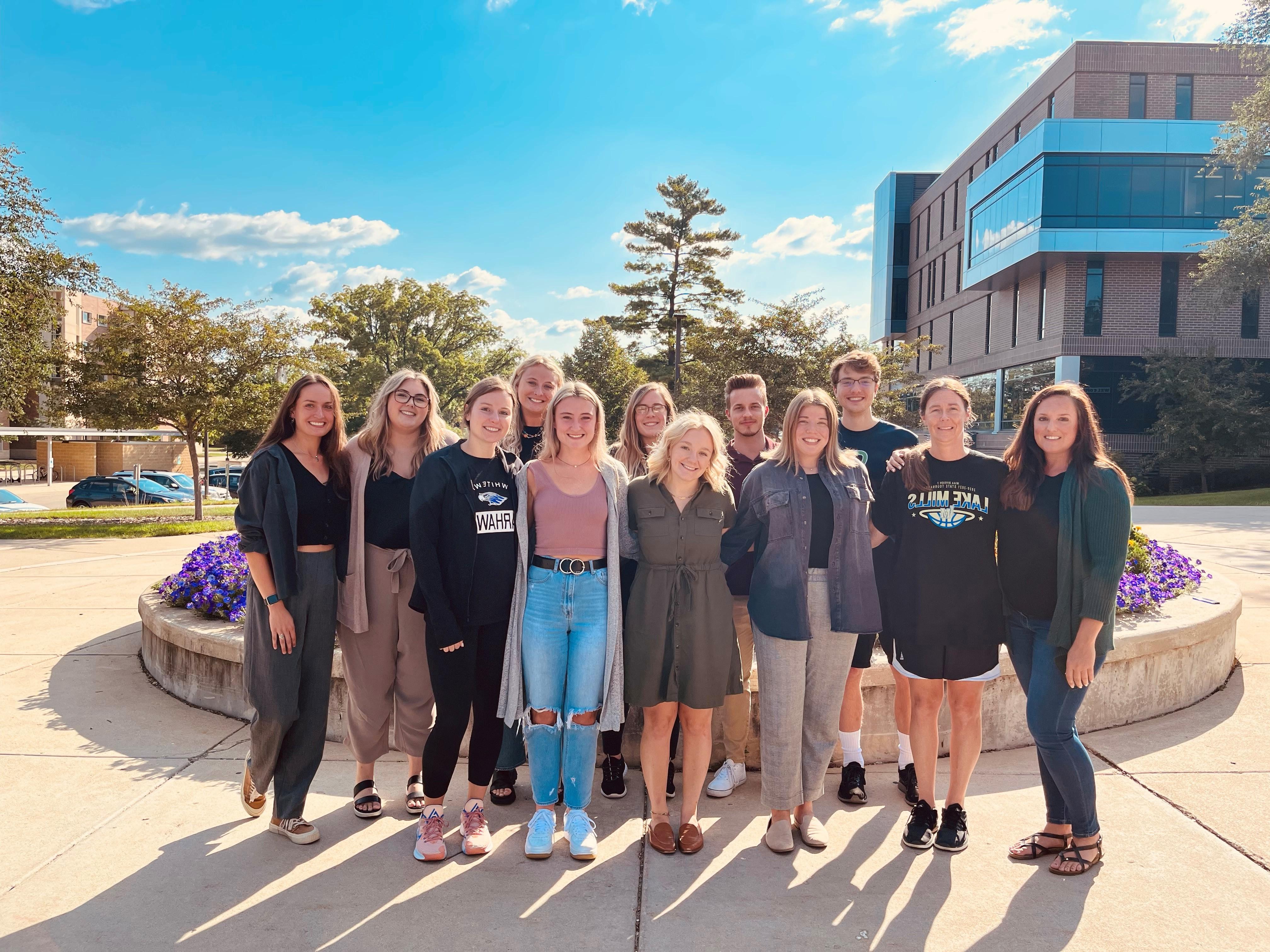 A group of people stand together in a row outdoors on campus.