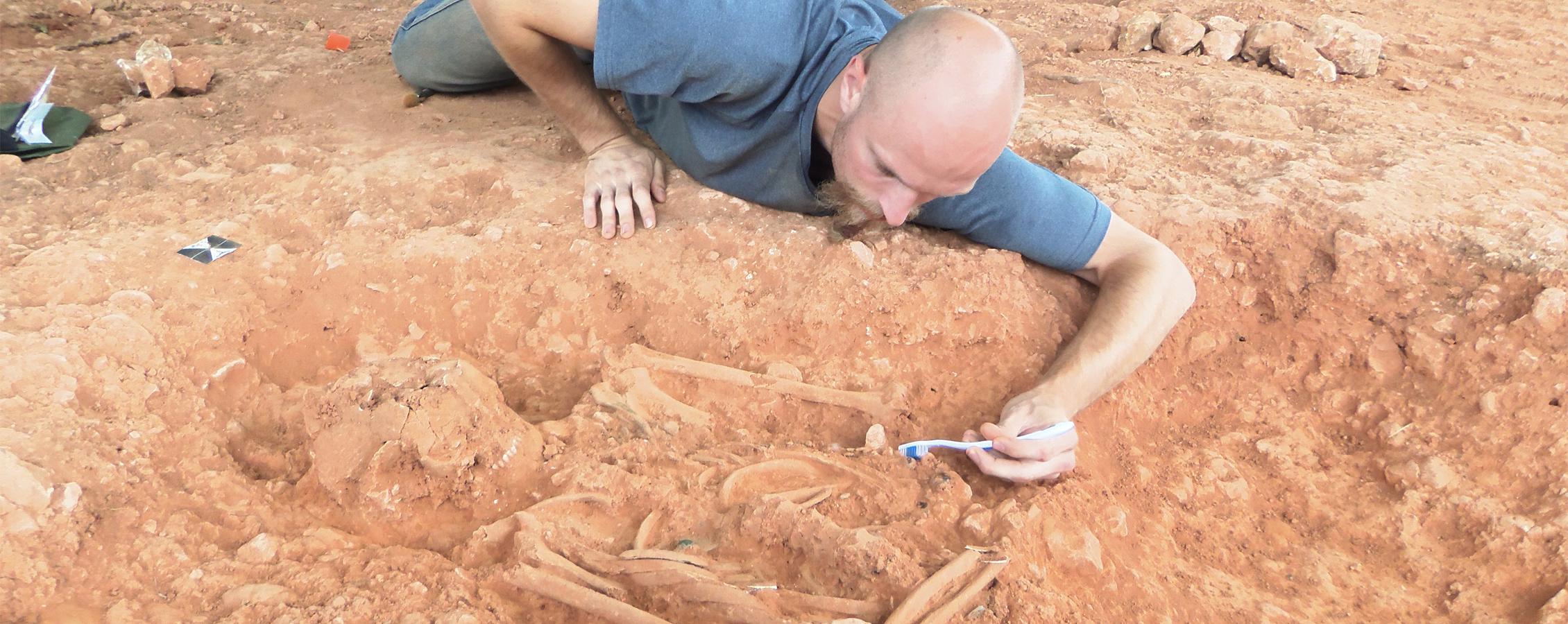 A student lays on the ground and uncovers a skeleton with a toothbrush.