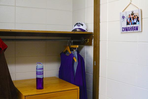 A wells hall dorm closet. There is a self in the closet with a dresser underneath it. There is a water bottle on top of the dresser. The shelf has a bar under it for hanging shirts, and there are multiple shirts hanging in this photo